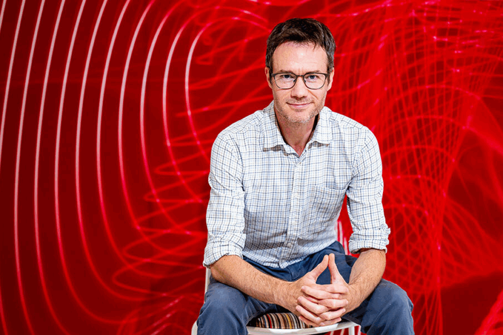 Geoff Boeing sits and poses for a photo in front of a red background.