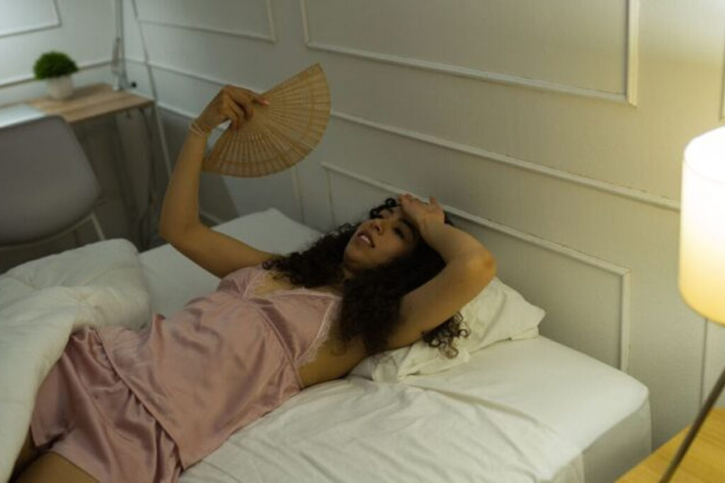 Photo shows young woman lying in bed awake fanning herself, unable to sleep.