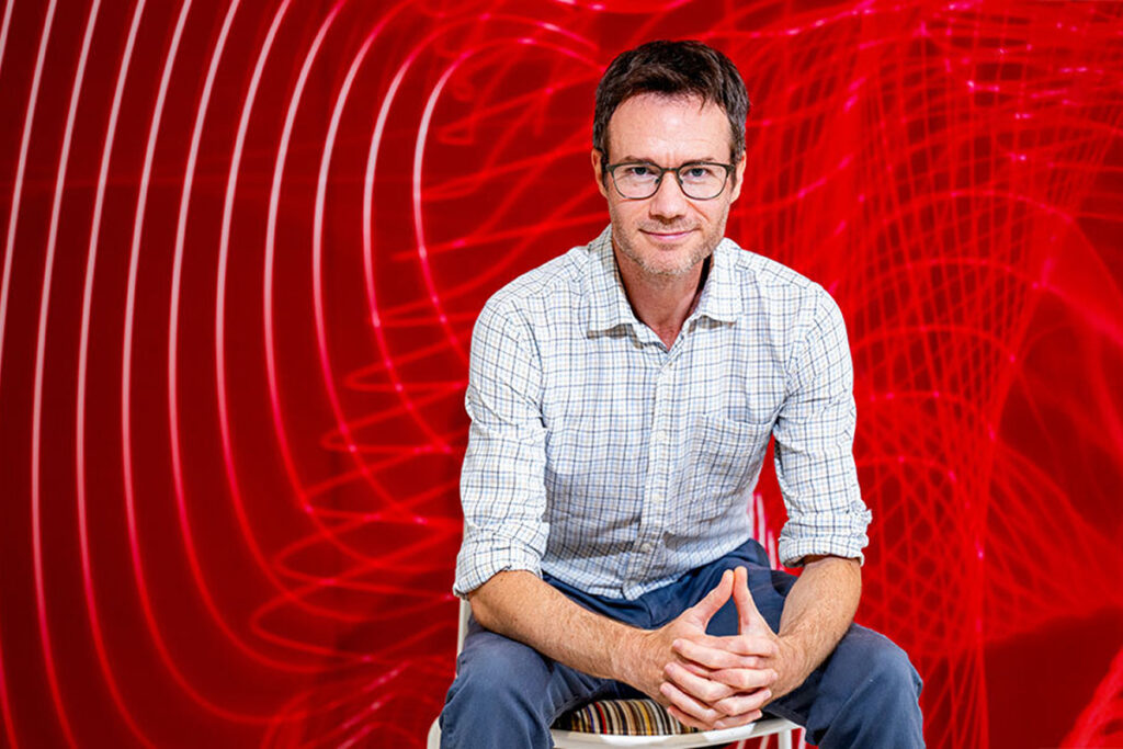 Geoff Boeing sits and poses for a photo in front of a red background.